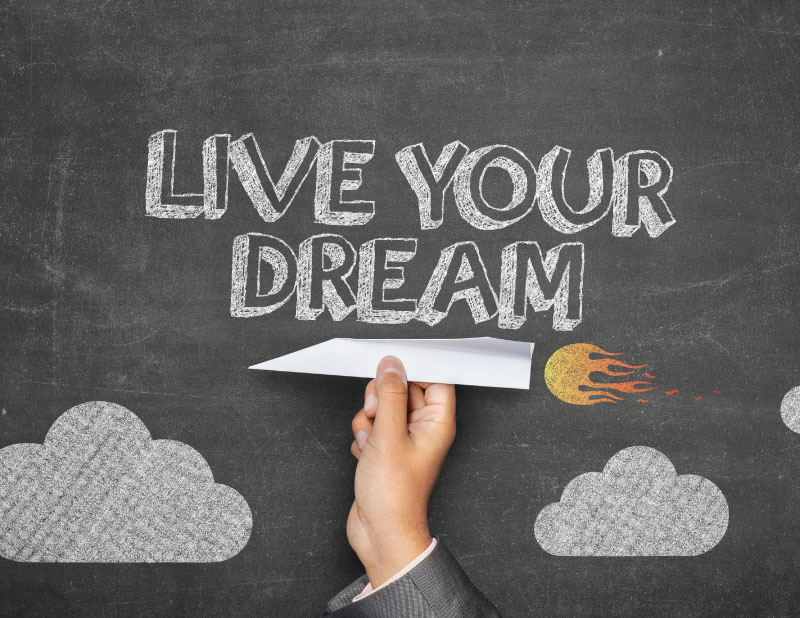hand holding a paper airplane in front of a chalk board with Live Your Dream written on it